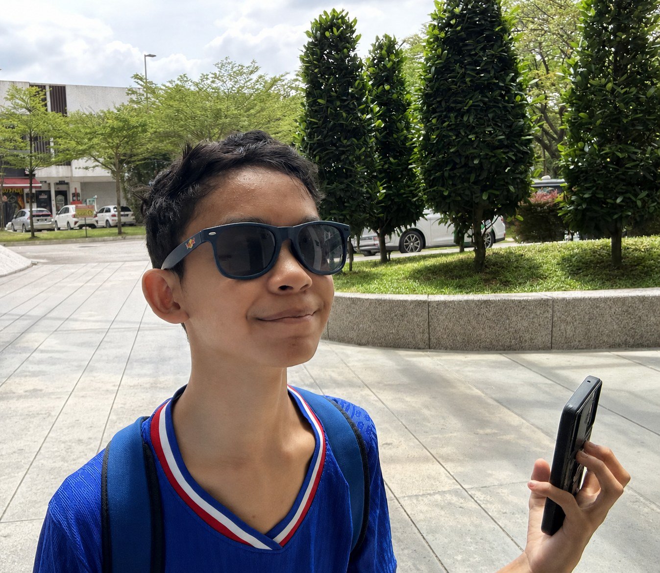 Raees, an autistic pianist, smiling outdoors in a blue jersey holding his phone