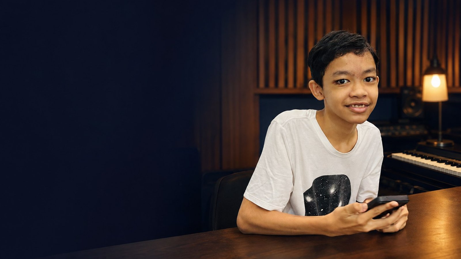 Raees, an autistic pianist, smiling and using his phone at a desk in his home recording studio